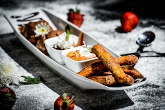 A white rectangular plate contains various desserts arranged aesthetically. On one side, churros coated with sugar and cinnamon are placed alongside small bowls filled with chocolate and caramel dips. Whipped cream with a mint leaf garnish adds a decorative touch. Whole strawberries are scattered on the surface dusted with powdered sugar, adding a vibrant contrast.