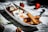 A white rectangular plate contains various desserts arranged aesthetically. On one side, churros coated with sugar and cinnamon are placed alongside small bowls filled with chocolate and caramel dips. Whipped cream with a mint leaf garnish adds a decorative touch. Whole strawberries are scattered on the surface dusted with powdered sugar, adding a vibrant contrast.