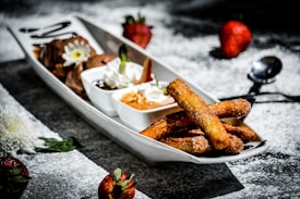 A white rectangular plate contains various desserts arranged aesthetically. On one side, churros coated with sugar and cinnamon are placed alongside small bowls filled with chocolate and caramel dips. Whipped cream with a mint leaf garnish adds a decorative touch. Whole strawberries are scattered on the surface dusted with powdered sugar, adding a vibrant contrast.