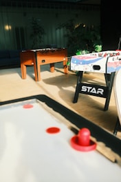 A room with air hockey and foosball tables. The air hockey table is in the foreground with a red paddle and puck. In the background, there is a wooden foosball table and another foosball table decorated with colorful stars. The room has potted plants and soft lighting contributes to a cozy atmosphere.