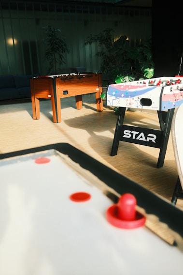 A room with air hockey and foosball tables. The air hockey table is in the foreground with a red paddle and puck. In the background, there is a wooden foosball table and another foosball table decorated with colorful stars. The room has potted plants and soft lighting contributes to a cozy atmosphere.