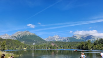 A serene landscape featuring a large lake surrounded by dense green forest with a backdrop of tall, rugged mountains under a clear blue sky. People are seen near the lake shore, some engaging in water activities. A triangular building is situated at the base of the mountains, partially obscured by trees.