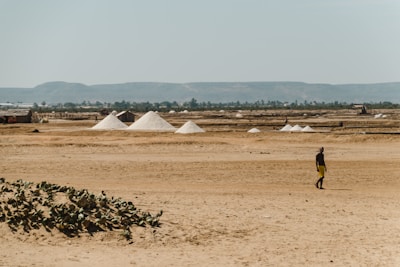 A vast, arid landscape with several large piles of white salt in the middle distance. The horizon features a range of low hills under a clear blue sky, and sparse vegetation can be seen on the barren ground. In the foreground, a person in yellow shorts walks across the sandy terrain.