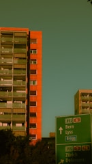 A tall orange apartment building stands prominently against a greenish sky. In the foreground, a green road sign directs to locations including Bern and Zürich. Trees are visible near the base of the building.
