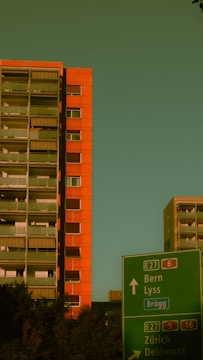 A tall orange apartment building stands prominently against a greenish sky. In the foreground, a green road sign directs to locations including Bern and Zürich. Trees are visible near the base of the building.