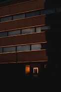 A building exterior is cast in deep shadow with horizontal lines of windows reflecting a warm light, likely from the setting or rising sun. The lower part of the image features illuminated signage with company logos partially visible, adding a contrast to the darkness.