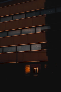 A building exterior is cast in deep shadow with horizontal lines of windows reflecting a warm light, likely from the setting or rising sun. The lower part of the image features illuminated signage with company logos partially visible, adding a contrast to the darkness.