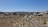 A desert landscape with sparse vegetation, showcasing a winding road cutting through the scene. Large boulders and rock formations line the background under a clear blue sky, and a few vehicles, including two motorcycles and a car, travel along the road.