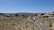A desert landscape with sparse vegetation, showcasing a winding road cutting through the scene. Large boulders and rock formations line the background under a clear blue sky, and a few vehicles, including two motorcycles and a car, travel along the road.