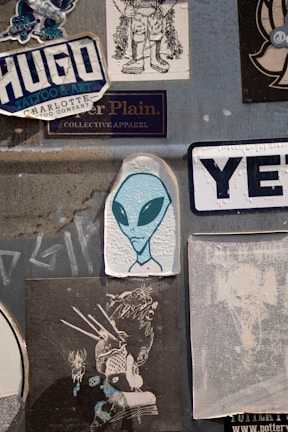 A collection of various stickers on a textured gray surface. Prominently features an illustration of an alien with large black eyes, surrounded by different graphic designs and brand logos. The stickers include stylized text and illustrations with urban and creative themes.