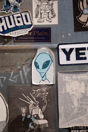 A collection of various stickers on a textured gray surface. Prominently features an illustration of an alien with large black eyes, surrounded by different graphic designs and brand logos. The stickers include stylized text and illustrations with urban and creative themes.