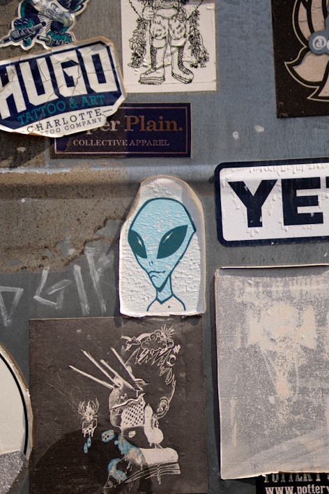 A collection of various stickers on a textured gray surface. Prominently features an illustration of an alien with large black eyes, surrounded by different graphic designs and brand logos. The stickers include stylized text and illustrations with urban and creative themes.