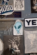 A collection of various stickers on a textured gray surface. Prominently features an illustration of an alien with large black eyes, surrounded by different graphic designs and brand logos. The stickers include stylized text and illustrations with urban and creative themes.