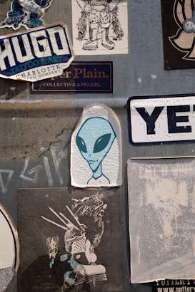 A collection of various stickers on a textured gray surface. Prominently features an illustration of an alien with large black eyes, surrounded by different graphic designs and brand logos. The stickers include stylized text and illustrations with urban and creative themes.