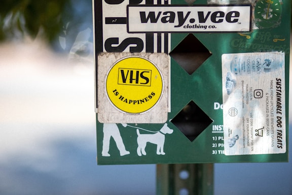 A collection of stickers and signs are attached to a surface, featuring a prominent yellow circular sticker with 'VHS is Happiness' written on it. Other elements include a white sticker advertising sustainable dog treats, a logo for a clothing company called 'way.vee,' and silhouettes of a person walking a dog.