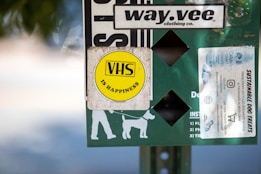 A collection of stickers and signs are attached to a surface, featuring a prominent yellow circular sticker with 'VHS is Happiness' written on it. Other elements include a white sticker advertising sustainable dog treats, a logo for a clothing company called 'way.vee,' and silhouettes of a person walking a dog.