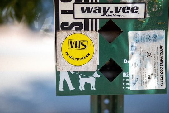 A collection of stickers and signs are attached to a surface, featuring a prominent yellow circular sticker with 'VHS is Happiness' written on it. Other elements include a white sticker advertising sustainable dog treats, a logo for a clothing company called 'way.vee,' and silhouettes of a person walking a dog.