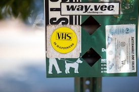 A collection of stickers and signs are attached to a surface, featuring a prominent yellow circular sticker with 'VHS is Happiness' written on it. Other elements include a white sticker advertising sustainable dog treats, a logo for a clothing company called 'way.vee,' and silhouettes of a person walking a dog.