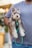 A small, fluffy dog with pointed ears and light brown and white fur is held by a person's arm. The dog wears a harness attached to a green and blue leash. The person is wearing a ribbed gray short-sleeve shirt and jeans. The background is slightly blurred, featuring hints of greenery and light tones.