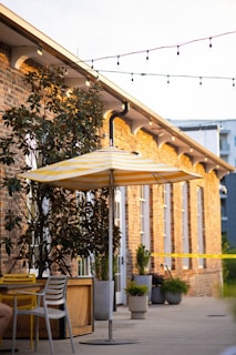 Outdoor seating area featuring a striped yellow and white umbrella and modern outdoor furniture, set against a brick building with several windows. Potted plants and string lights add to the ambiance, and a wet paint caution tape is visible in the background.