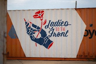 A mural painted on a cargo container featuring a stylized hand holding a red flower. Beside the image, bold text reads 'Ladies to the Front' in a combination of blue and red colors. Both the text and the artwork create a bold and empowering statement.