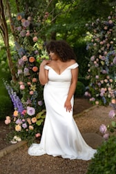 A woman wearing an elegant off-shoulder white wedding gown stands in a lush garden setting, surrounded by an archway made of various colorful flowers and greenery. The floral arrangement includes roses and other blooms in shades of purple, pink, cream, and peach. She gazes thoughtfully to the side, with her hand raised to her neckline.