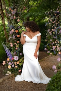 A woman wearing an elegant off-shoulder white wedding gown stands in a lush garden setting, surrounded by an archway made of various colorful flowers and greenery. The floral arrangement includes roses and other blooms in shades of purple, pink, cream, and peach. She gazes thoughtfully to the side, with her hand raised to her neckline.