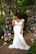 A woman wearing an elegant off-shoulder white wedding gown stands in a lush garden setting, surrounded by an archway made of various colorful flowers and greenery. The floral arrangement includes roses and other blooms in shades of purple, pink, cream, and peach. She gazes thoughtfully to the side, with her hand raised to her neckline.