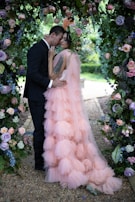 A couple dressed in formal attire stands closely together under an arch adorned with lush greenery and colorful flowers. The woman is wearing a flowing, tiered pink dress with a flower crown, while the man is in a black suit. The scene is beautifully set in a garden with flowers in shades of purple, pink, and white surrounding them.