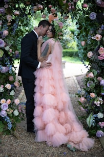 A couple dressed in formal attire stands closely together under an arch adorned with lush greenery and colorful flowers. The woman is wearing a flowing, tiered pink dress with a flower crown, while the man is in a black suit. The scene is beautifully set in a garden with flowers in shades of purple, pink, and white surrounding them.