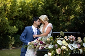 A couple is sharing an intimate moment, with the groom in a blue suit and the bride in a white dress adorned with floral designs. They are surrounded by lush green foliage and a beautiful arrangement of pastel-colored flowers.