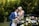A couple is sharing an intimate moment, with the groom in a blue suit and the bride in a white dress adorned with floral designs. They are surrounded by lush green foliage and a beautiful arrangement of pastel-colored flowers.