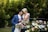 A couple is sharing an intimate moment, with the groom in a blue suit and the bride in a white dress adorned with floral designs. They are surrounded by lush green foliage and a beautiful arrangement of pastel-colored flowers.