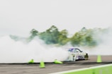A car is drifting on a track surrounded by thick smoke, with bright green traffic cones scattered around. The car, decorated with vibrant decals, creates an impression of speed and control. Trees are visible in the blurred background, adding to the dynamic scene.