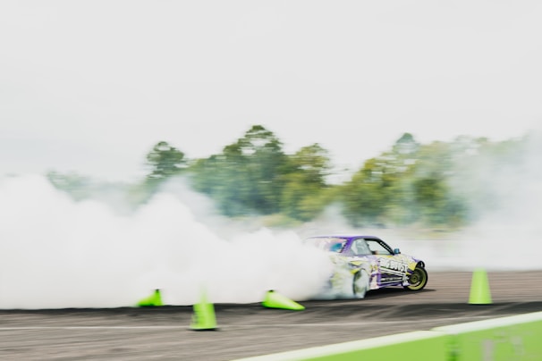A car is drifting on a track surrounded by thick smoke, with bright green traffic cones scattered around. The car, decorated with vibrant decals, creates an impression of speed and control. Trees are visible in the blurred background, adding to the dynamic scene.