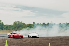 Two cars are drifting on a track surrounded by smoke and greenery in the background. One car is red, while the other is white. They are maneuvering closely around traffic cones on an open asphalt area.