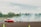 Two cars are drifting on a track surrounded by smoke and greenery in the background. One car is red, while the other is white. They are maneuvering closely around traffic cones on an open asphalt area.