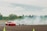 Two cars are drifting on a track surrounded by smoke and greenery in the background. One car is red, while the other is white. They are maneuvering closely around traffic cones on an open asphalt area.