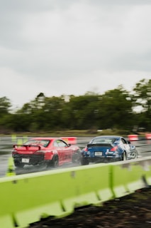 Two sports cars, a red one and a blue one, are racing on a wet track. The surroundings include greenery and road safety barriers, with motion blur indicating high speed. There are some orange and white traffic cones or barriers in the background.