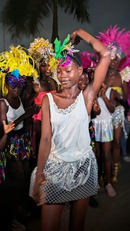 A group of people, mainly young women, are dressed in colorful and elaborate costumes, featuring vibrant feathered headdresses and glittery skirts. The central figure is dancing, captured mid-movement, with a joyful expression. The background is filled with more dancers in similarly festive attire, creating a lively and celebratory atmosphere.