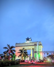 A monumental arch structure illuminated with colorful lights, featuring a prominent black star on top. The inscription 'AD 1957 Freedom and Justice' is visible. Palm trees and decorative lighting create a vibrant atmosphere, while people are gathered around the structure.