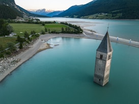 A picturesque scene featuring a submerged bell tower rising from the calm, turquoise waters of a lake. Surrounded by lush green fields and dense forests, the landscape extends towards distant snow-capped mountains. The serene setting is captured during what appears to be dusk or dawn, with soft lighting enhancing the tranquil atmosphere.
