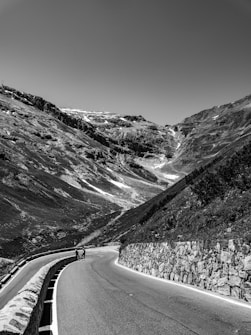 A winding mountain road bordered by stone walls stretches into the distance surrounded by rugged terrain. Two cyclists are visible, riding up the incline at a distance, adding a human element to the vast landscape. The sky is clear and the monochrome effect emphasizes the texture of the rocks and road.