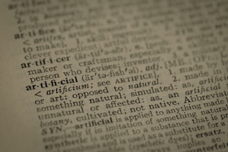 A close-up view of a dictionary page highlighting the word 'artificial' along with its pronunciation, part of speech, and definition. The text surrounding 'artificial' is printed in a traditional serif font on a slightly textured paper.