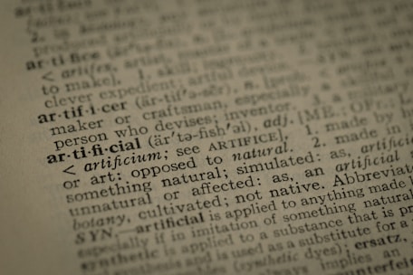 A close-up view of a dictionary page highlighting the word 'artificial' along with its pronunciation, part of speech, and definition. The text surrounding 'artificial' is printed in a traditional serif font on a slightly textured paper.