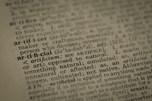 A close-up view of a dictionary page highlighting the word 'artificial' along with its pronunciation, part of speech, and definition. The text surrounding 'artificial' is printed in a traditional serif font on a slightly textured paper.