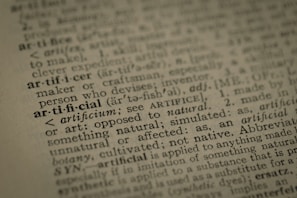 A close-up view of a dictionary page highlighting the word 'artificial' along with its pronunciation, part of speech, and definition. The text surrounding 'artificial' is printed in a traditional serif font on a slightly textured paper.