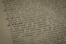 A close-up view of a dictionary page highlighting the word 'artificial' along with its pronunciation, part of speech, and definition. The text surrounding 'artificial' is printed in a traditional serif font on a slightly textured paper.