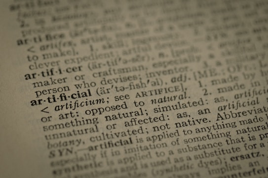 A close-up view of a dictionary page highlighting the word 'artificial' along with its pronunciation, part of speech, and definition. The text surrounding 'artificial' is printed in a traditional serif font on a slightly textured paper.