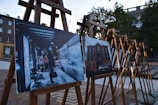 Several easels are set up outdoors, each displaying photographs. The image in the foreground shows a nighttime scene at a bus stop with a person in a long coat and a bright vest, holding a bag. The second image depicts a brick building with a car parked in front.